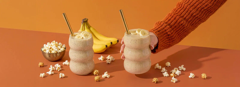 Two MaxiMeal banana milkshakes with fresh bananas and popcorn on an orange background