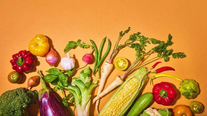 Fresh vegetables and fruit arranged on an orange background, illustrating the whole, nutrient-dense food choices that modern food environments make harder to prioritise for MaxiMeal blog