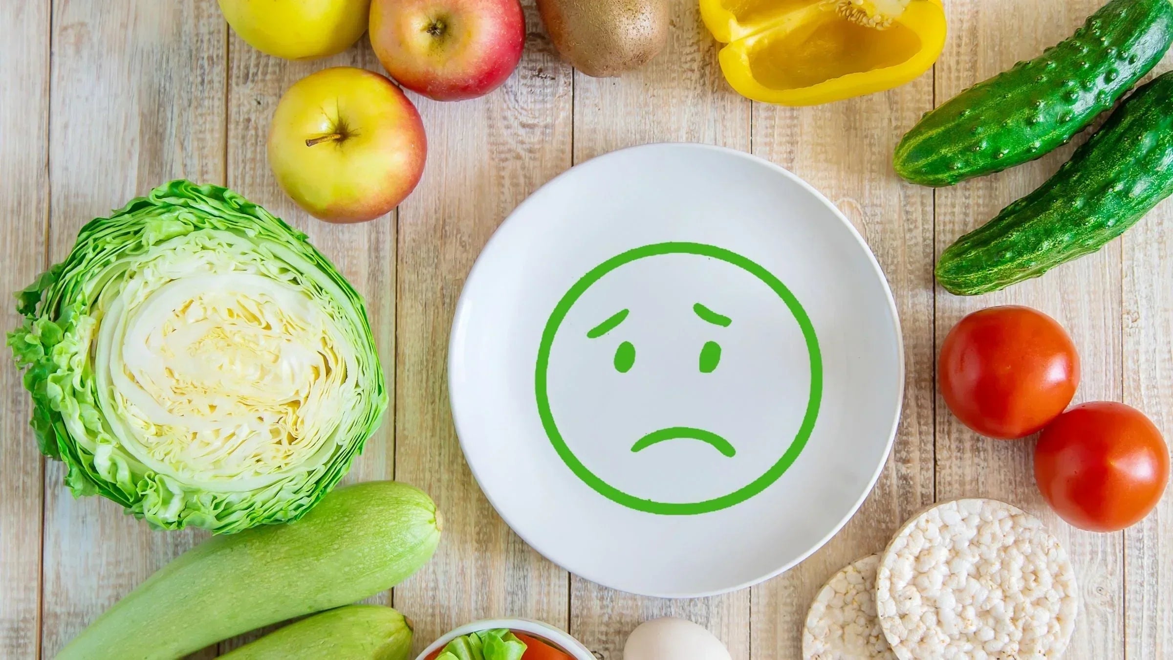 A sad face drawn on an empty plate surrounded by fresh vegetables and fruit, illustrating the mealtime frustration parents of fussy eaters often face for MaxiMeal blog